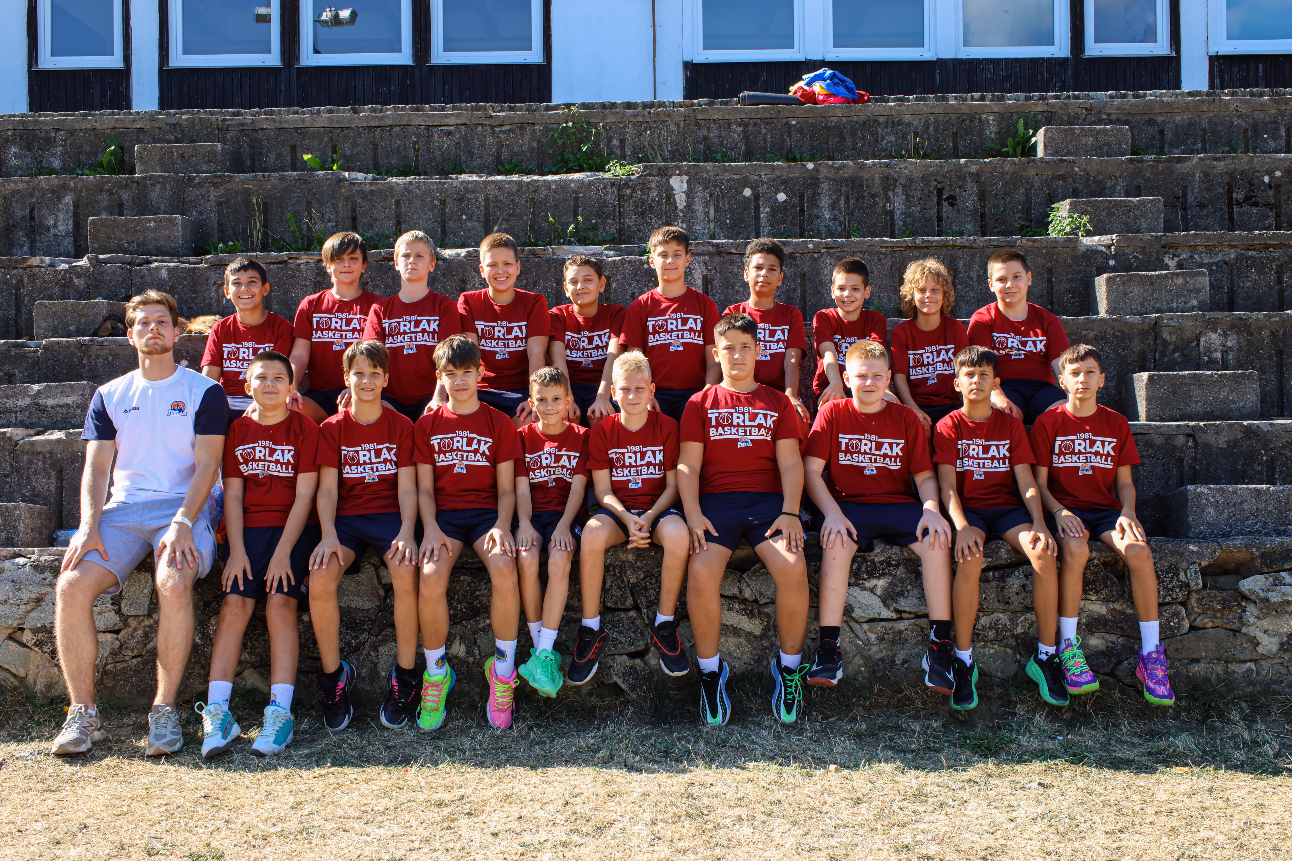 Group photo of a youth basketball team and their coach sitting on stone steps outdoors. The coach wears a white t-shirt and the players wear matching maroon t-shirts with 'Torlak Basketball' printed on them, paired with dark shorts and sneakers.